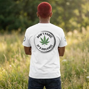 May include: White t-shirt with a circular graphic on the back featuring a green cannabis leaf and the text "DON'T PANIC IT'S ORGANIC". The shirt is worn by a person with red hair, standing in a field of tall grass.