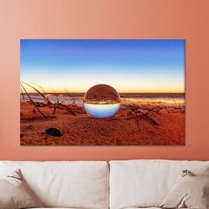 May include: A glass sphere lens sits on a sandy beach, reflecting a blue sky and a golden sunset over the ocean.