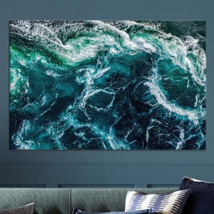 May include: A rectangular canvas print showing an aerial view of ocean waves. The water shades from deep teal to turquoise, with white foamy crests. The artwork is displayed on a blue wall above a sofa with cushions.