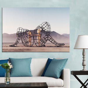 May include: A canvas print featuring two wireframe sculptures of seated figures, back-to-back, on a desert landscape. The sculptures have a grid-like design, with a light-colored figure inside each. Mountains are visible in the background.