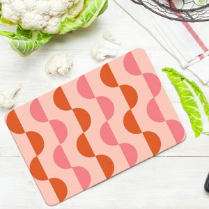 May include: A rectangular cutting board with a pink background and a repeating pattern of orange and pink semi-circles. A stainless steel knife with a black handle is next to the board. Cauliflower and green leaves are also in the image.