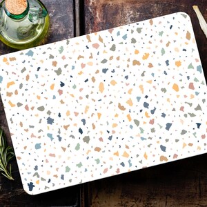 Könnte beinhalten: Ein rechteckiges weißes Schneidebrett mit einem Terrazzo-Muster in verschiedenen Farben. Zutaten wie Rosmarin, Knoblauch und Olivenöl umgeben das Brett.