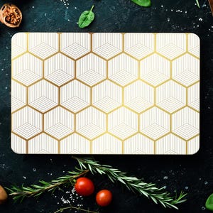 Può includere: Tagliere rettangolare bianco con un motivo geometrico dorato a nido d'ape. La tavola è circondata da ingredienti come pomodori, noci ed erbe aromatiche, suggerendo un tema culinario. Il design aggiunge un tocco di eleganza.