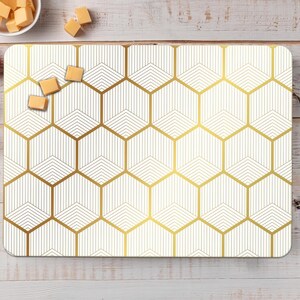 May include: Rectangular white cutting board with a gold geometric honeycomb pattern. The board is surrounded by fresh tomatoes, rosemary, cheese cubes, bay leaves, and peppercorns on a light wood surface.