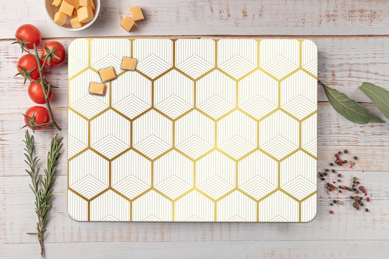 May include: Rectangular white cutting board with a gold geometric honeycomb pattern. The board is surrounded by fresh tomatoes, rosemary, cheese cubes, bay leaves, and peppercorns on a light wood surface.