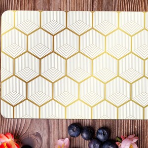 May include: A rectangular placemat with a geometric gold honeycomb pattern on a cream background. The placemat is surrounded by food items, including toast with jam, croissants, and blueberries, on a wooden surface.