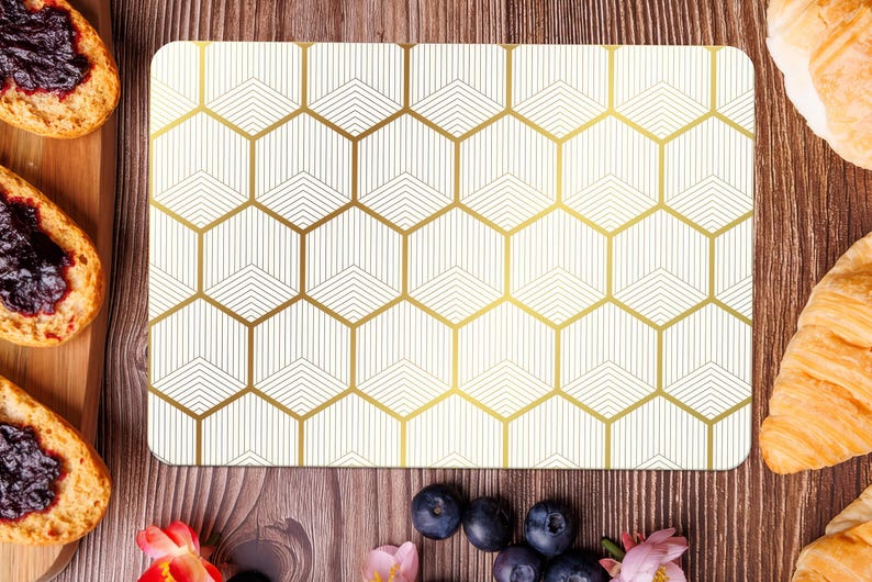 May include: A rectangular placemat with a geometric gold honeycomb pattern on a cream background. The placemat is surrounded by food items, including toast with jam, croissants, and blueberries, on a wooden surface.