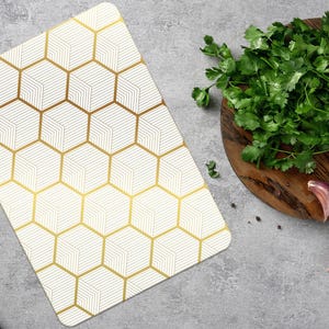 May include: A white cutting board with a gold geometric pattern sits next to a wooden cutting board with fresh herbs, spices, and garlic. The cutting board has a gold honeycomb design.