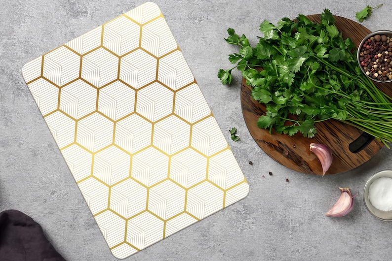 May include: A white cutting board with a gold geometric pattern sits next to a wooden cutting board with fresh herbs, spices, and garlic. The cutting board has a gold honeycomb design.