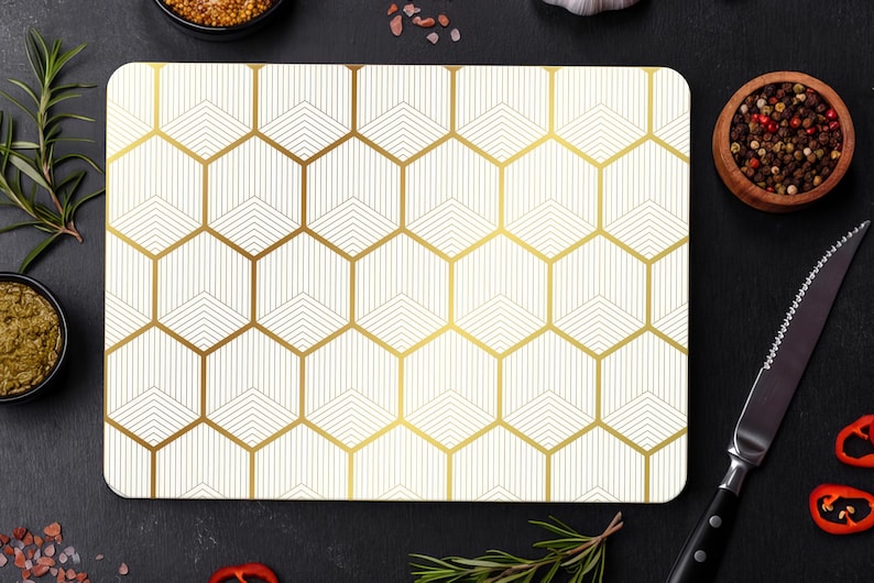 May include: Rectangular cutting board with a geometric gold and white pattern. The board is surrounded by ingredients like spices, herbs, and a knife, suggesting a culinary theme. The design features a repeating hexagon pattern.