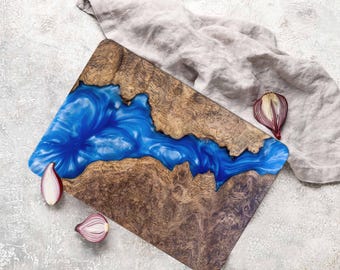 May include: A wooden chopping board with a blue epoxy resin inlay that resembles a river. The board has a natural wood grain pattern and is a unique and decorative piece.