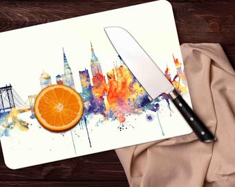 May include: A white cutting board with a watercolor painting of the New York City skyline. The painting features a bridge, buildings, and a splash of orange, yellow, and blue colors. A silver knife with a black handle is on the cutting board.