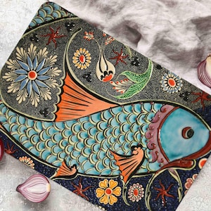 Puede incluir: Un plato de servir de cerámica con un diseño de pez azul y naranja. El pez tiene escamas y está rodeado de patrones florales en tonos de verde, amarillo y rojo. El plato está sobre una superficie blanca.