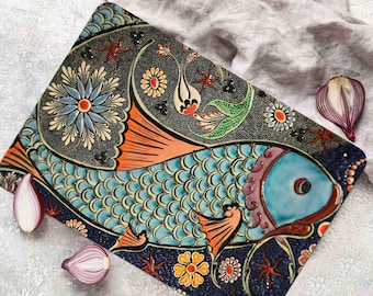 May include: A ceramic serving platter with a blue and orange fish design. The fish has scales and is surrounded by floral patterns in shades of green, yellow, and red. The platter is on a white surface.