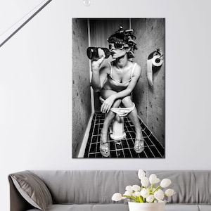 May include: Black and white wall art print depicting a person wearing sunglasses and hair curlers, sitting on a toilet and drinking from a bottle. The image is set in a bathroom with a toilet roll holder.