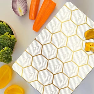 May include: Overhead shot of a white and gold geometric pattern cutting board with fresh vegetables. Broccoli in a bowl, yellow bell peppers, carrots, and red onion are arranged around the board.