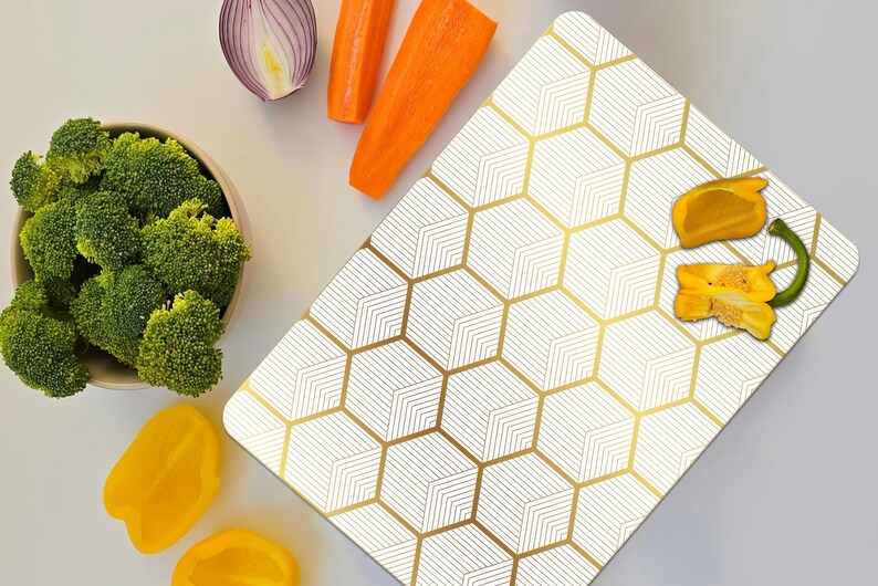 May include: Overhead shot of a white and gold geometric pattern cutting board with fresh vegetables. Broccoli in a bowl, yellow bell peppers, carrots, and red onion are arranged around the board.
