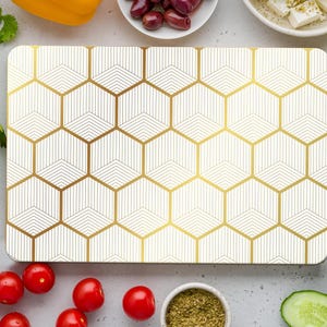 May include: A rectangular cutting board with a geometric gold and white pattern. The board is surrounded by fresh vegetables, including red tomatoes, cucumbers, and bell peppers. Olives, feta cheese, and spices are also present.