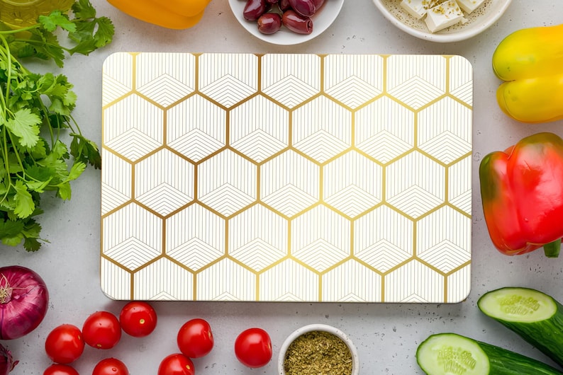 May include: A rectangular cutting board with a geometric gold and white pattern. The board is surrounded by fresh vegetables, including red tomatoes, cucumbers, and bell peppers. Olives, feta cheese, and spices are also present.