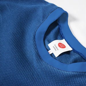 Op de afbeelding: Close-up van een blauw getextureerd T-shirt met ronde hals en een contrasterende blauwe kraag. Een wit label met Japanse tekst en een rood-wit ontwerp is aan de kraag bevestigd.