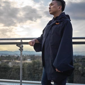 May include: A navy blue hooded coat with wide sleeves and a wooden toggle closure. The person is wearing dark blue jeans and standing on a rooftop with a glass railing, overlooking a cityscape under a cloudy sky.