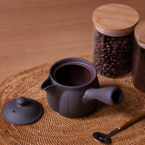 May include: A black ceramic teapot with a lid and a handle, sitting on a woven mat with two glass jars filled with coffee beans and coffee grounds. A silver spoon is also on the mat.