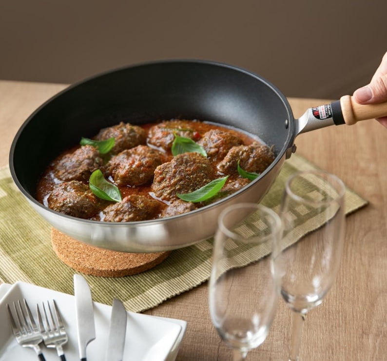 Pu&ograve; includere: Una padella d'argento con manico di legno, piena di salsa rossa e cibo simile a polpette ricoperto di erbe verdi.