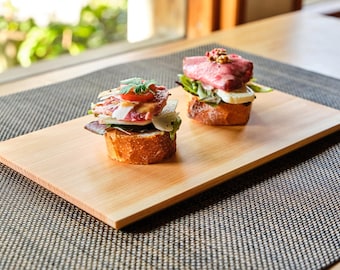 May include: Two open-faced sandwiches on a rectangular wooden serving board. The sandwiches are on toasted bread and topped with various ingredients, including meat, vegetables, and herbs. The board sits on a textured gray placemat.