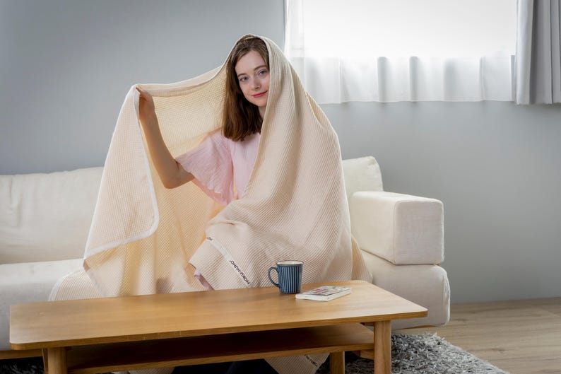 May include: A woman sits on a white couch, wrapped in a cream-colored waffle weave throw blanket. She is holding the blanket up to her face and smiling. A blue ceramic mug sits on a wooden table in front of her.