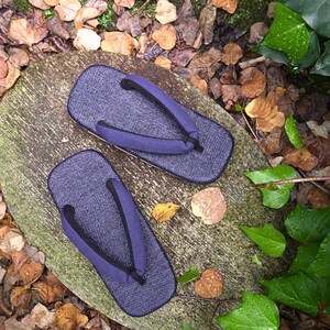 May include: A pair of dark blue traditional Japanese sandals, known as geta, with black straps. The sandals are sitting on a wooden surface with fallen leaves scattered around.