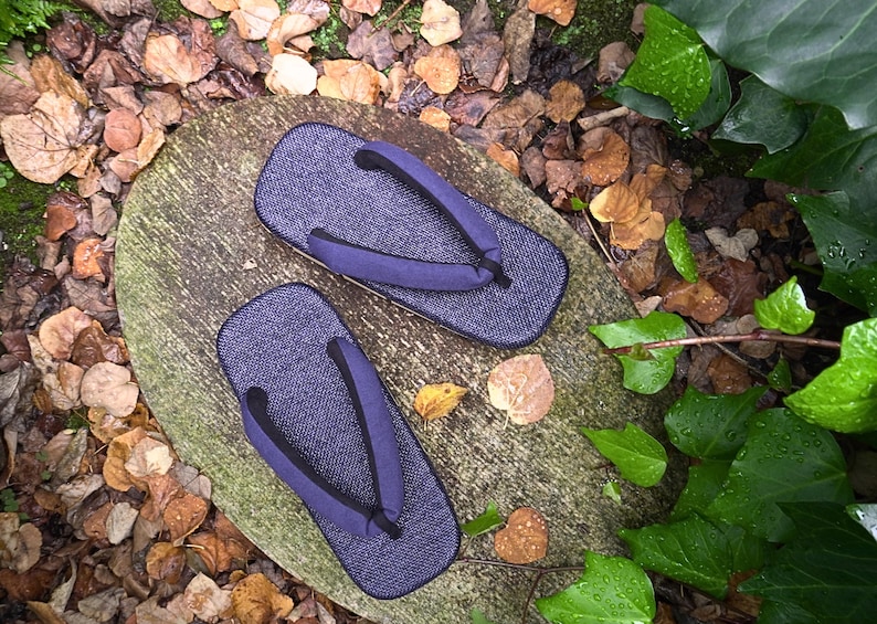 May include: A pair of dark blue traditional Japanese sandals, known as geta, with black straps. The sandals are sitting on a wooden surface with fallen leaves scattered around.