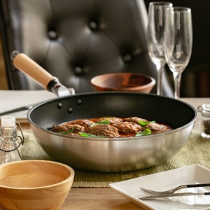 Pu&ograve; includere: Una padella d'argento con manico di legno, piena di polpette in salsa di pomodoro, guarnite con erbe fresche. La padella &egrave; appoggiata su un sottopentola verde su un tavolo di legno.