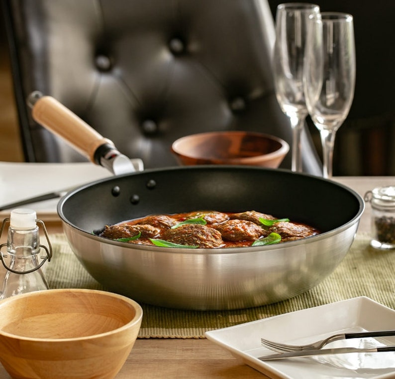 Pu&ograve; includere: Una padella d'argento con manico di legno, piena di polpette in salsa di pomodoro, guarnite con erbe fresche. La padella &egrave; appoggiata su un sottopentola verde su un tavolo di legno.