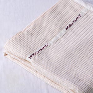 May include: A cream-colored waffle weave throw blanket with a white label that says "MOKU-NUNO" in burgundy lettering.