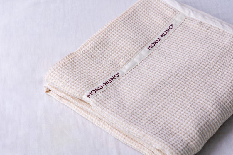 May include: A cream-colored waffle weave throw blanket with a white label that says "MOKU-NUNO" in burgundy lettering.