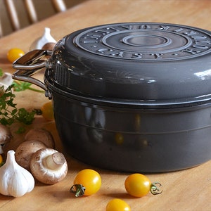 Puede incluir: Una olla holandesa de hierro fundido gris con tapa y dos asas. La olla está sobre una mesa de madera con tomates amarillos, champiñones y dientes de ajo.