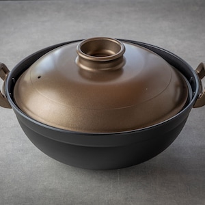 Puede incluir: Una olla redonda negra y dorada con dos asas y una tapa. La olla tiene una superficie texturizada y la tapa tiene un pequeño pomo en la parte superior.