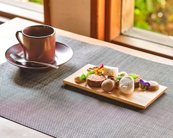 May include: A brown ceramic coffee cup and saucer with a spoon, alongside a wooden serving board with various pastries and garnishes. The scene is set on a textured gray placemat near a window.