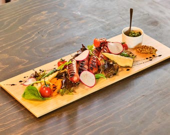 May include: A wooden serving board displays a gourmet meal. Grilled sausages, tomatoes, radishes, and greens are arranged with a small cup of sauce and a side of bread. The food is drizzled with a dark sauce.