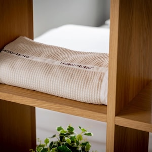 May include: A white waffle weave throw blanket with the text "MOKU-NUNO" printed on it, resting on a wooden shelf.