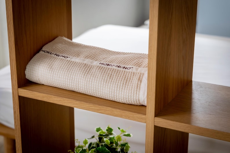 May include: A white waffle weave throw blanket with the text "MOKU-NUNO" printed on it, resting on a wooden shelf.