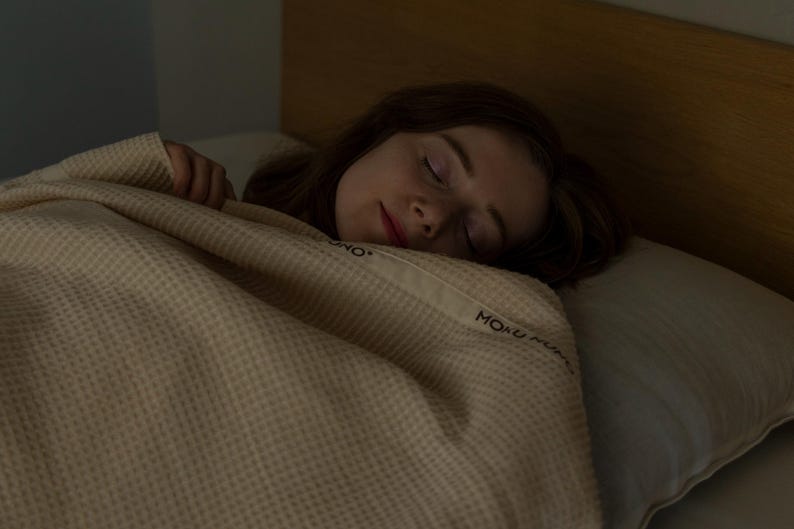 May include: A person sleeping under a beige waffle weave blanket with the text "MONO MONO" embroidered on it.
