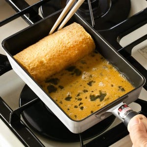 May include: A square-shaped, non-stick frying pan with a black handle is being used to make a rolled omelette. The omelette is being rolled with chopsticks. The pan is on a gas hob.