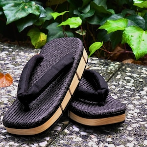May include: A pair of black and brown traditional Japanese sandals, known as geta, with a woven fabric top and wooden base. The sandals are sitting on a stone surface.