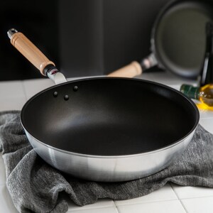 Pu&ograve; includere: Una padella argentata e nera con manico in legno. La padella &egrave; appoggiata su un asciugamano grigio.