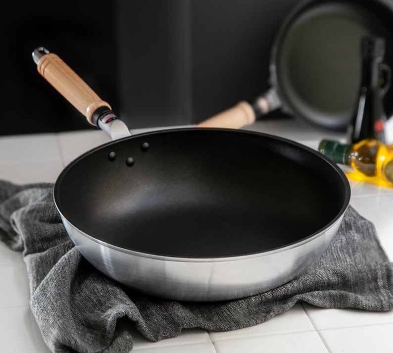 Pu&ograve; includere: Una padella argentata e nera con manico in legno. La padella &egrave; appoggiata su un asciugamano grigio.