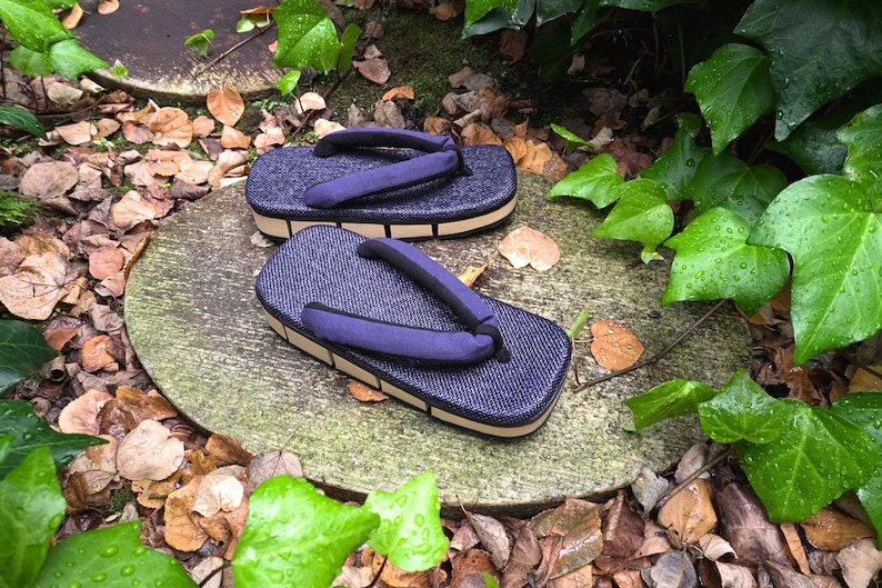 May include: Two pairs of blue and beige Japanese sandals, called geta, on a stone surface with green leaves