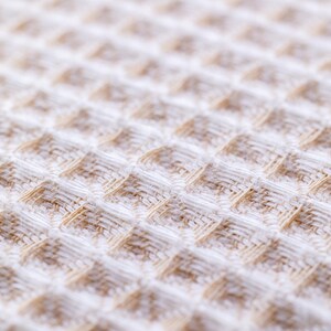 May include: Close-up of a white waffle weave fabric with a subtle brown pattern. The fabric has a soft, textured surface.