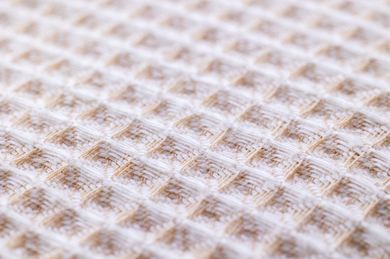 May include: Close-up of a white waffle weave fabric with a subtle brown pattern. The fabric has a soft, textured surface.