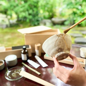 Pode incluir: Uma tigela de chá de cerâmica branca com um esmalte craquelado e manchas marrons. A tigela está sendo segurada por uma mão, e um batedor de madeira está descansando na borda. A tigela é cercada por outros suprimentos para a cerimônia do chá, incluindo uma caixa de madeira, um pequeno frasco e uma colher de madeira.